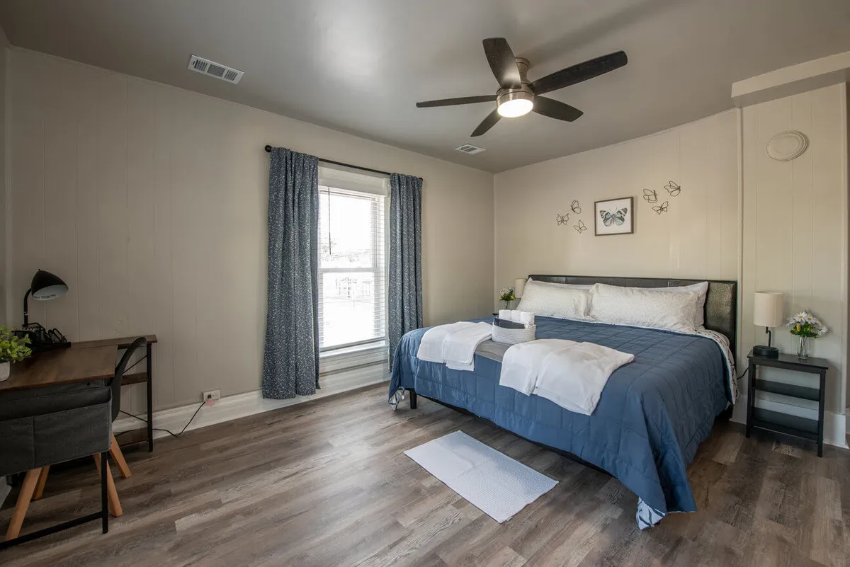 A peaceful bedroom scene with a cozy bed, a desk beside a window, and decorative wall art. The space is bathed in gentle natural light, providing a soothing environment for relaxing, reading, or enjoying quiet moments of solitude.