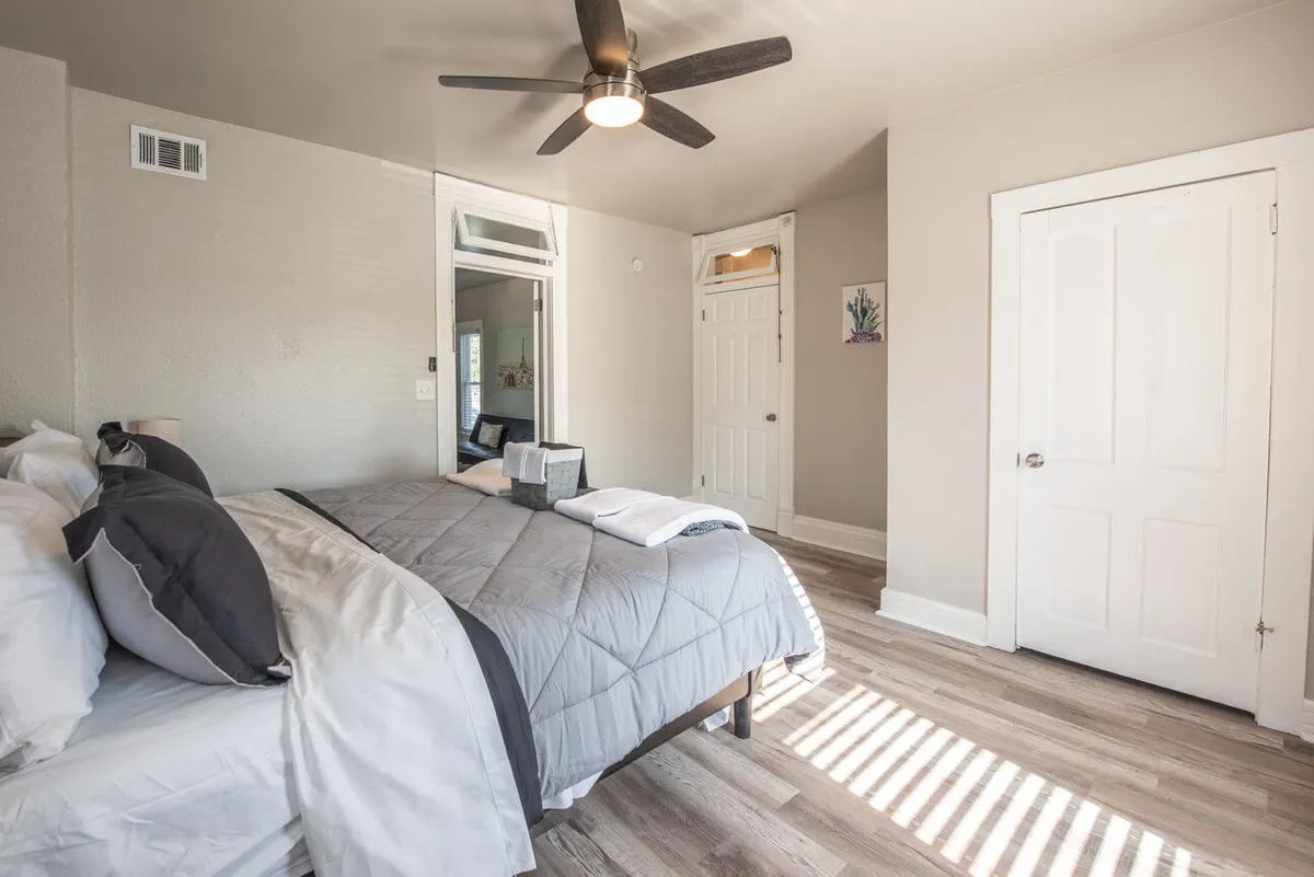 A tranquil bedroom scene with a bed dressed in gray bedding, complemented by a ceiling fan and wooden flooring. Decorative artwork adds a touch of personality to the serene setting, and ample sunlight streams in, illuminating the space and creating a welcoming atmosphere.