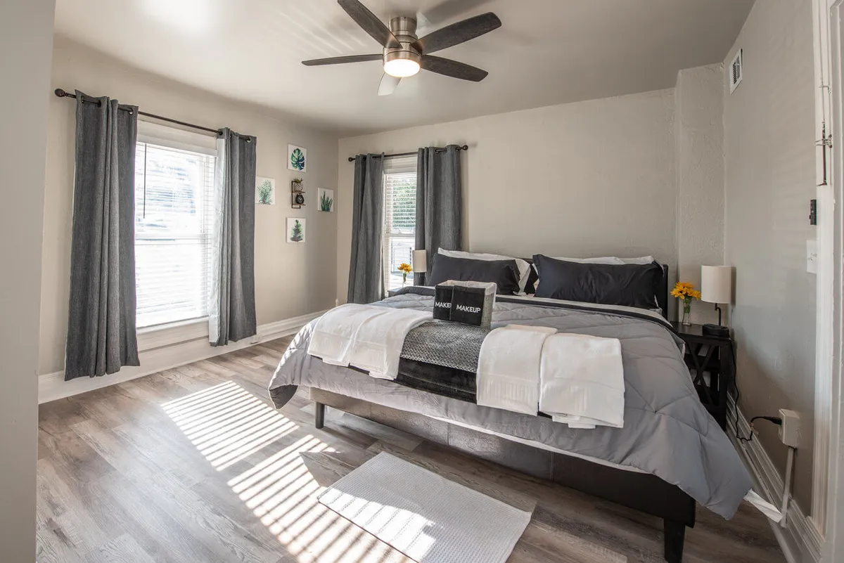 A tranquil bedroom featuring a comfortable bed with gray bedding, decorative wall art above the headboard, and soft natural light streaming through generous windows. The ceiling fan and cozy blankets add to the restful atmosphere, creating a perfect spot for relaxation and rejuvenation.