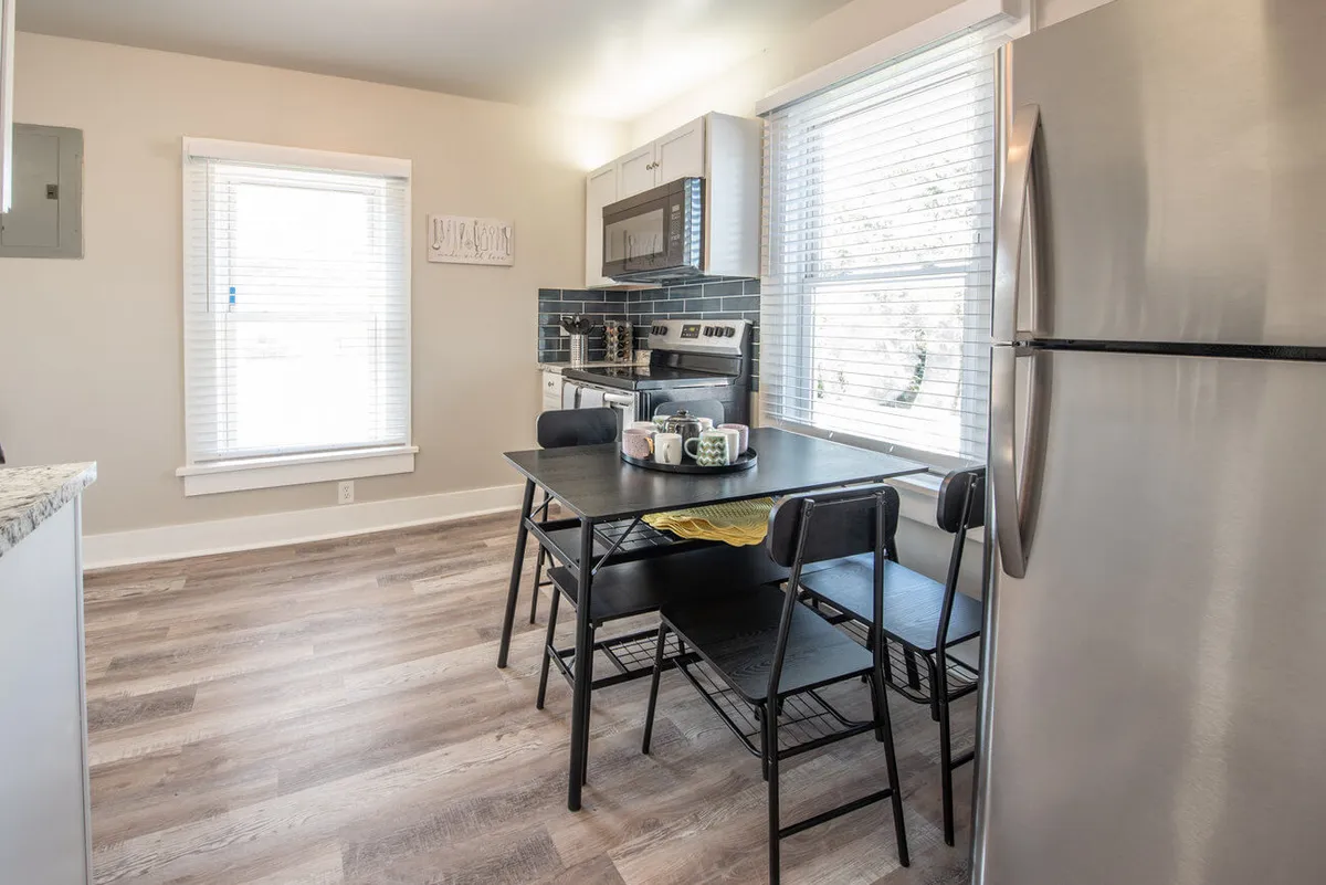 A bright and inviting kitchen scene that showcases a dining table adjacent to well-appointed kitchen appliances, all bathed in natural light from generous windows. The setup encourages socializing and shared meals, reinforcing the kitchen's function as a hub for cooking, dining, and connecting with others.