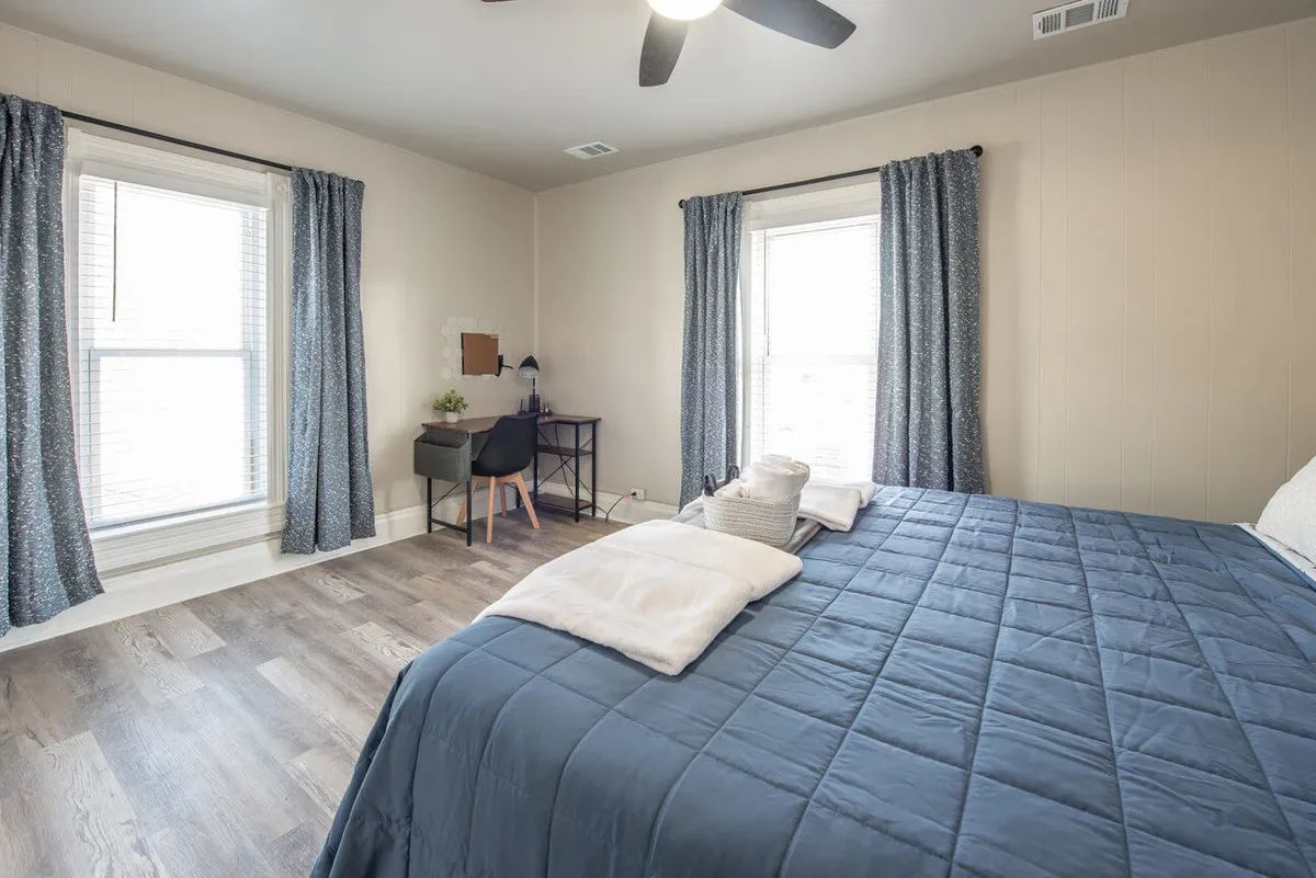 A serene bedroom with a bed dressed in a blue quilt, a desk and chair for reading or working, and large windows adorned with curtains that let in plenty of natural light. The space exudes comfort and offers a welcoming retreat for relaxation.